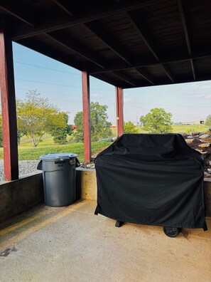 A covered barbecue grill and a trash bin under a wooden structure with a view of a grassy field and trees.