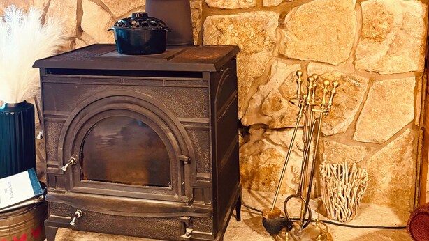A stone wall with a wood-burning stove, a metal pot on top, and a broom leaning against the wall.