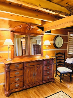 A wooden cabin interior with a large wooden dresser, two wall-mounted lamps, a clock, and a chair.