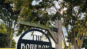 A circular sign with 'THE BOURBON LODGE' text, mounted on a wooden post, surrounded by grass and trees.