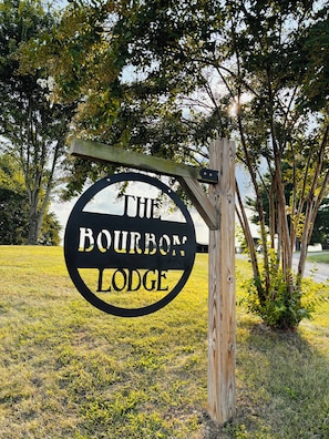 A circular sign with 'THE BOURBON LODGE' text, mounted on a wooden post, surrounded by grass and trees.