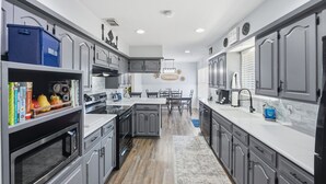 A modern kitchen with grey cabinets, a white countertop, and a central island. There is a blue storage bin labeled "BREAD" on the left, and a microwave below it. The kitchen has a wooden floor and a dining area with a table and chairs in the background. - Quiet Farm, Gameroom, Firepit 2 Mi to McKinney (McKinney)