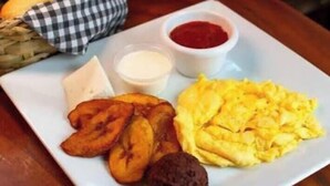 Une assiette contenant des bananes plantains frites, des œufs brouillés, une tranche de pain et deux petits récipients de condiments.