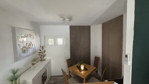 Un comedor con una mesa, sillas, un florero con flores, una lámpara de pared y una ventana con persianas.
