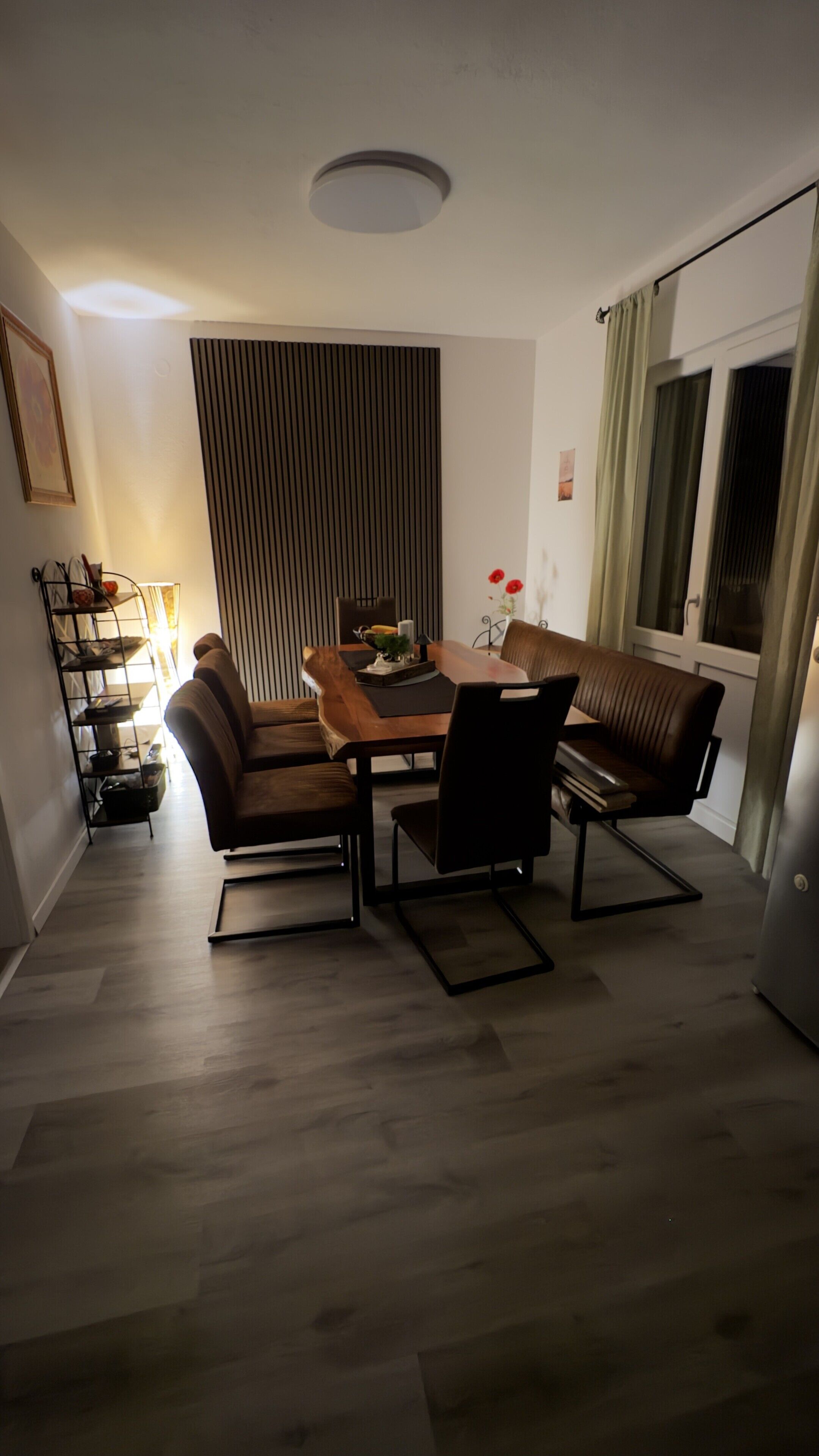 En matplats med ett tr äbord, fyra stolar, en soffa, en taklampa, ett fönster med gardiner och en vägghylla med böcker och dekorativa föremål.