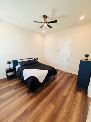 Une chambre à coucher comprenant un lit, une table de chevet, un bureau, un ventilateur de plafond et une porte.