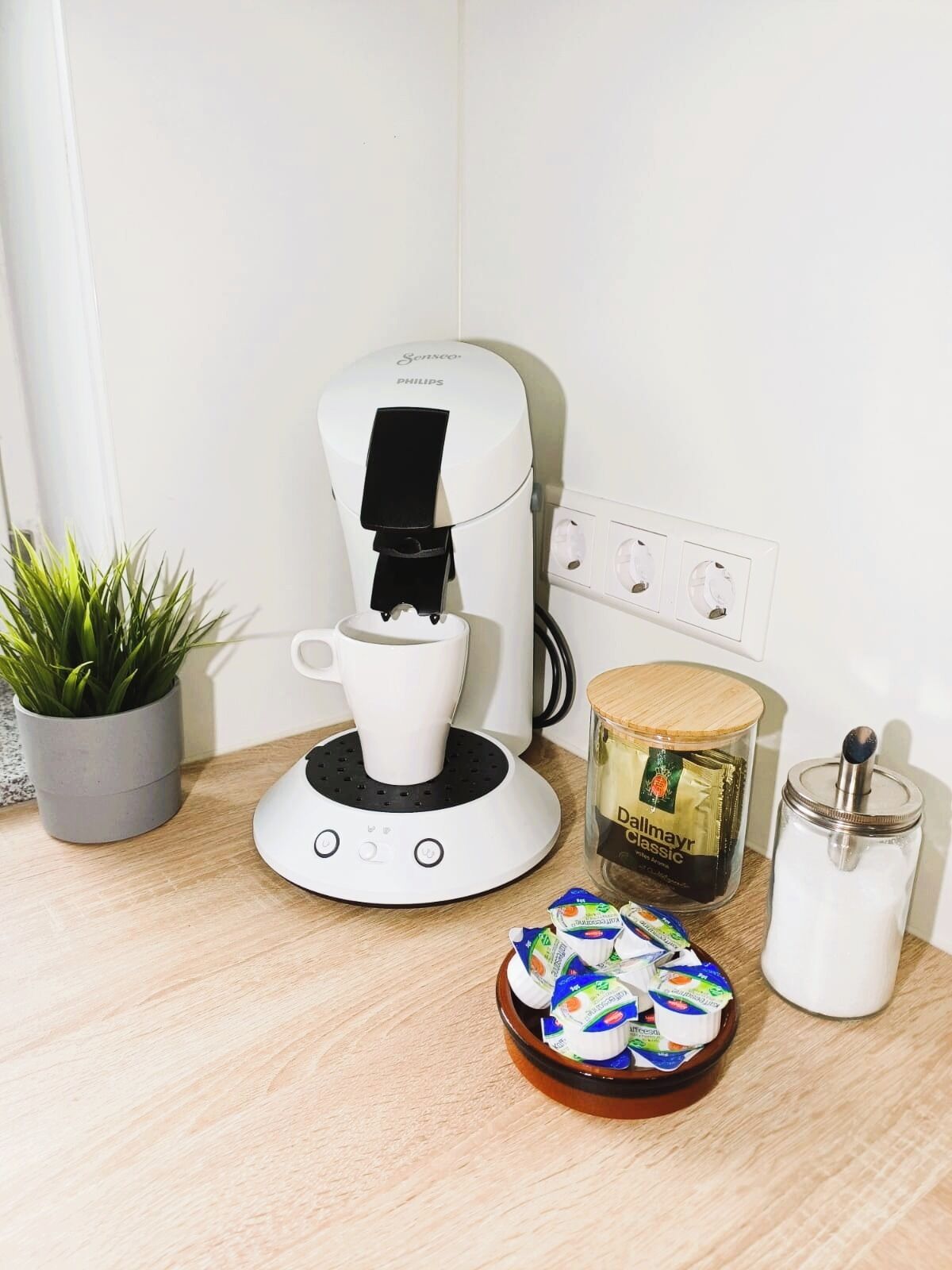 Een koffiezetapparaat, een plant, een pot koffie, een suikerpot en een bakje yoghurt op een houten oppervlak.