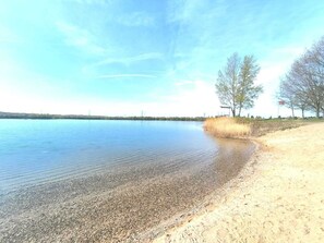 Un llac tranquil amb riba pedregosa, cel serè i alguns arbres dispersos.