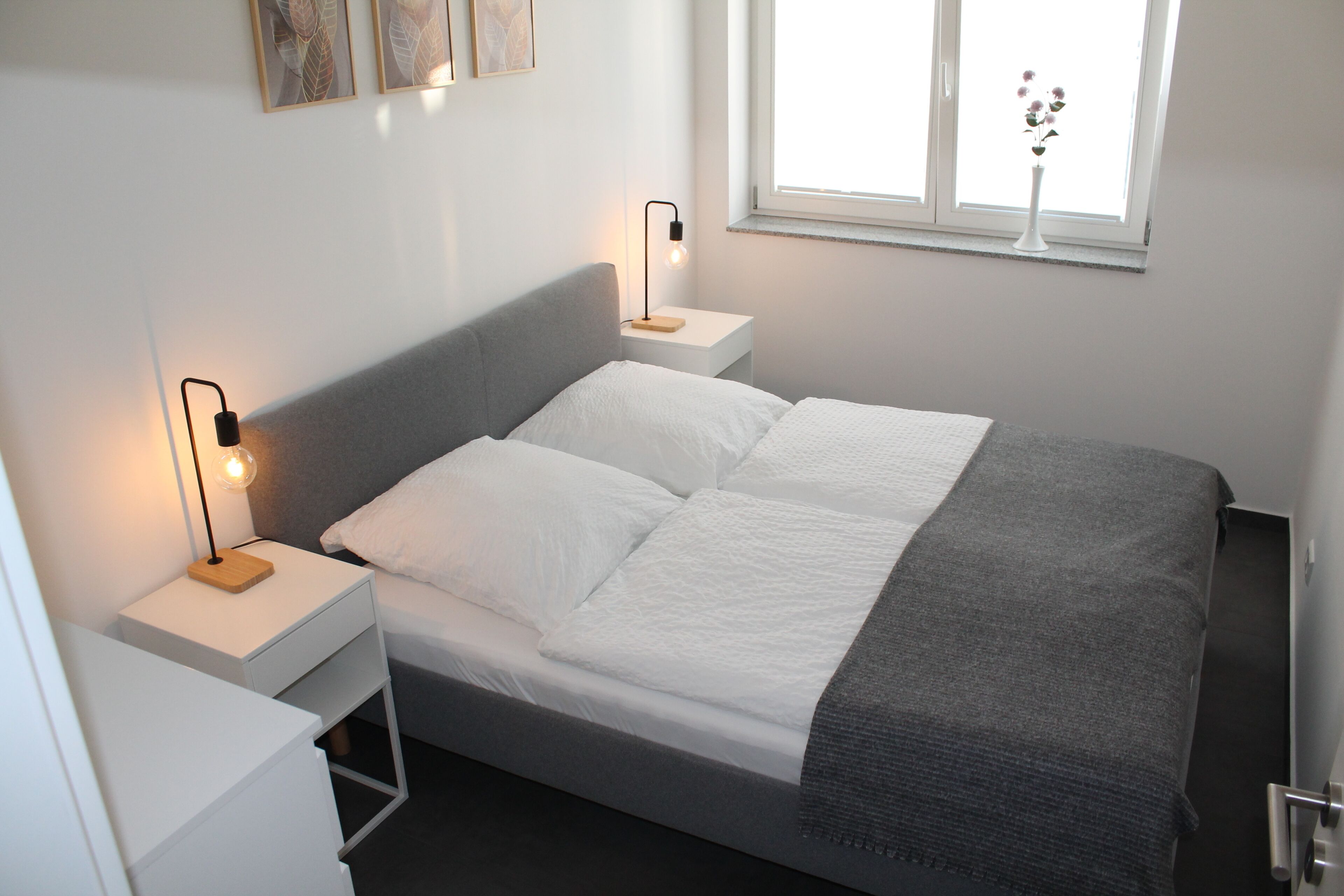 Un lit bien fait, avec du linge de lit blanc et un plaid gris, deux tables de chevet avec des lampes noires, un vase blanc orné de fleurs sur le rebord de la fenêtre, et un mur blanc décoré d’œuvres d’art encadrées.