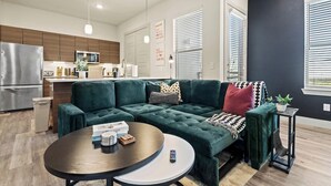 Ein modernes Wohnzimmer mit einem grünen Ecksofa, einem runden Couchtisch und einer Küchenzeile mit Holzschränken und Edelstahlgeräten.