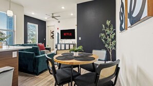 Ein modernes Wohnzimmer mit dunkler Akzentwand, einem Flachbildfernseher, einem Essbereich mit rundem Tisch und Stühlen sowie einer gemütlichen Sitzecke mit Sofa und Kissen.