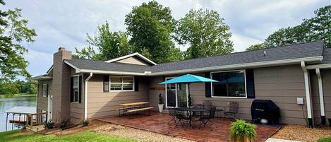 A house with a patio, a table, chairs, a grill, and a view of a lake.