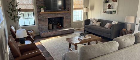 Una sala de estar con chimenea de ladrillo, un televisor de pantalla plana, un sofá gris, un sillón de cuero marrón y una mesa de centro de madera.