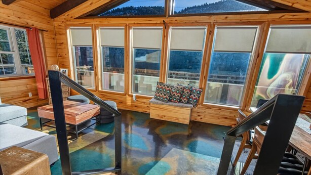 L’intérieur d’un chalet en bois, doté de grandes fenêtres, d’un escalier en bois et d’un banc agrémenté de coussins colorés.