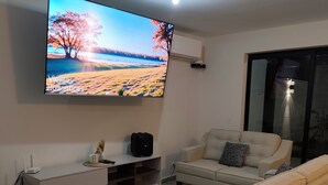 Un soggiorno moderno con una televisione a schermo piatto montata al muro, un divano color beige, una poltrona bianca e un tavolino da caffè in legno.
