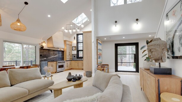 A spacious living area with a sofa, coffee table, and a dining table. A kitchen with modern appliances and wooden cabinets is visible in the background. - A Modern, Ultra Chic 5-bedroom Beach Home in Charming East Hampton! (East Hampton)