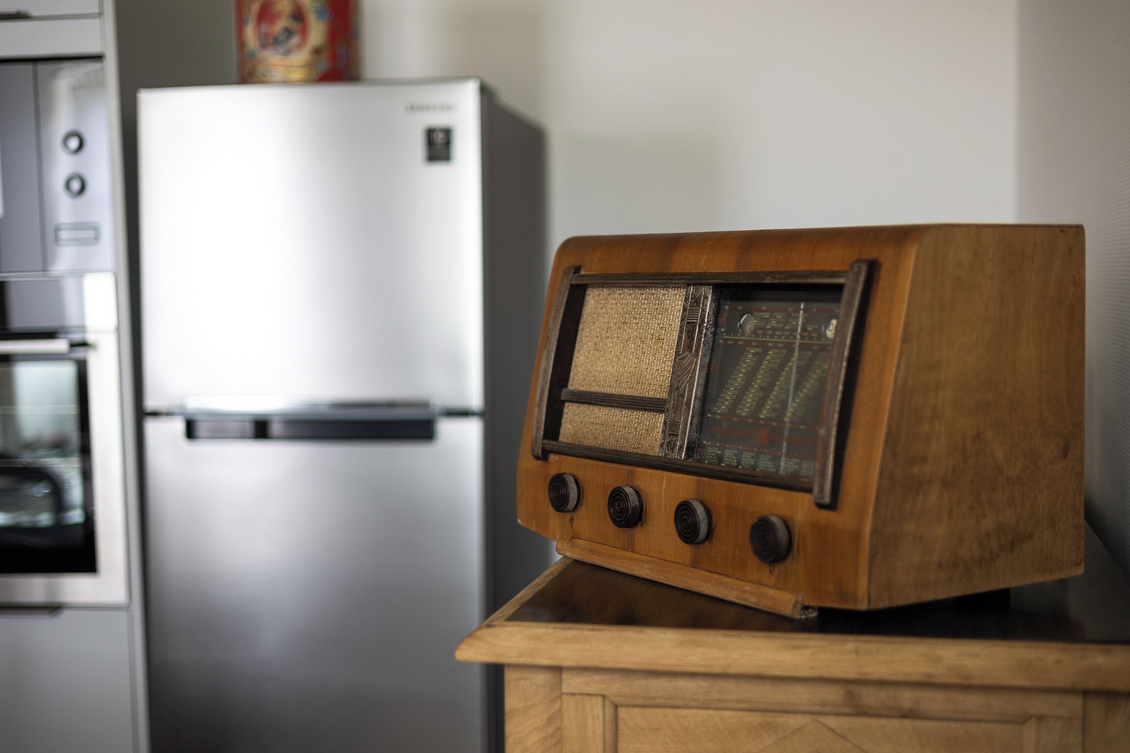 A wooden radio on a wooden cabinet in a kitchen with a stainless steel refrigerator in the background.