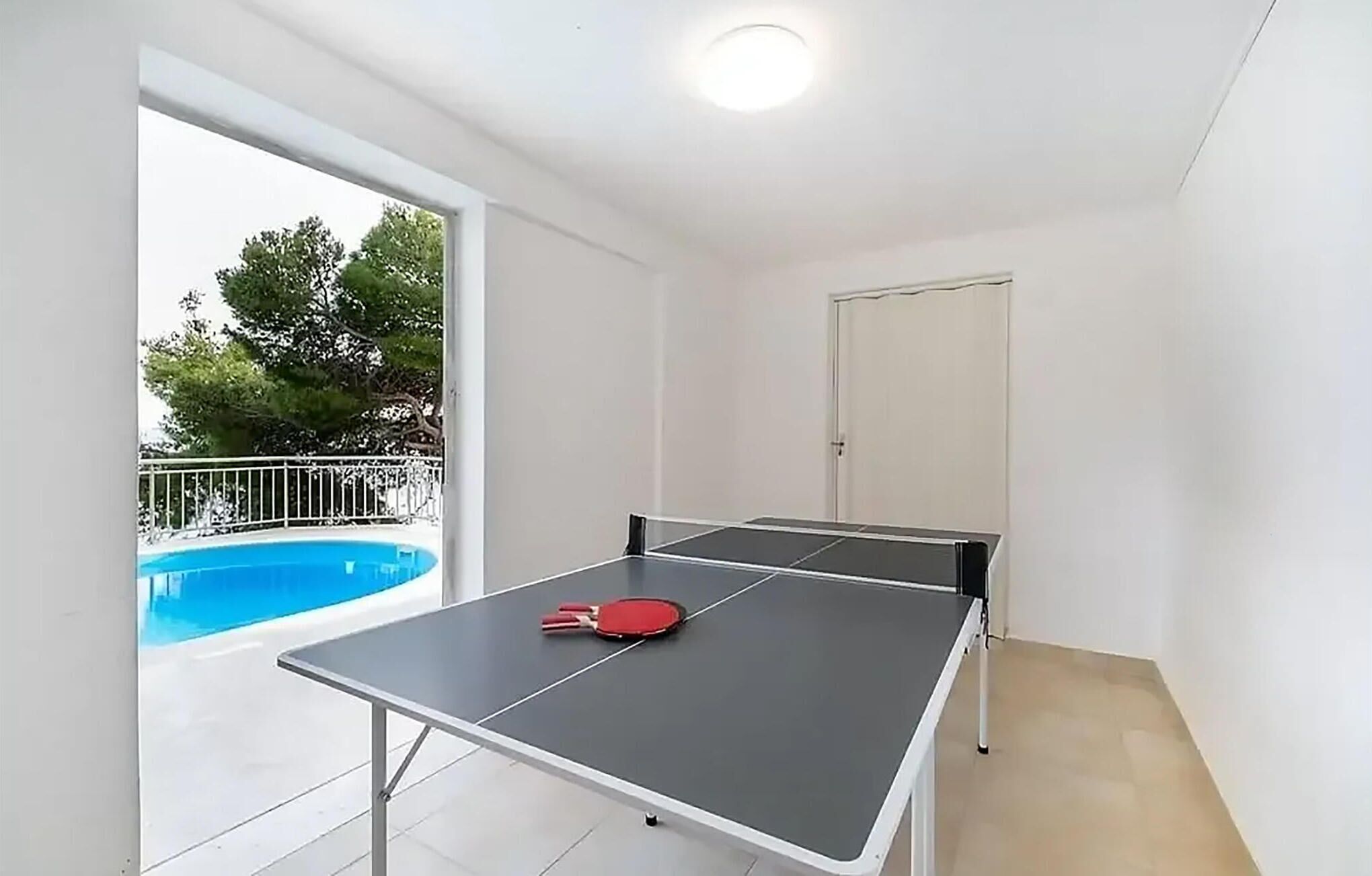 A room with a ping pong table, a door, and a view of a swimming pool.