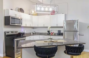 A modern kitchen with white cabinets, a stainless steel refrigerator, a microwave, an oven, and a granite countertop. There are two dark chairs at the island.