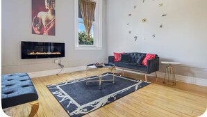 Een woonkamer met een zwarte bank, een open haard, een klok aan de muur en een raam met gordijnen.