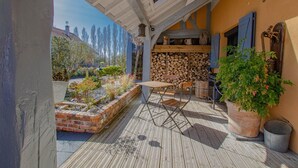 Eine überdachte Terrasse mit einem Holztisch und -stühlen, einem Ziegelpflanztopf mit Pflanzen und einem Holzstoß.