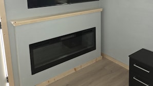 Una habitación con un televisor montado en la pared, una chimenea debajo y un mueble negro pequeño a la derecha.