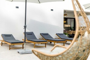 A row of sun loungers with cushions, a hanging chair, and a wall-mounted umbrella.