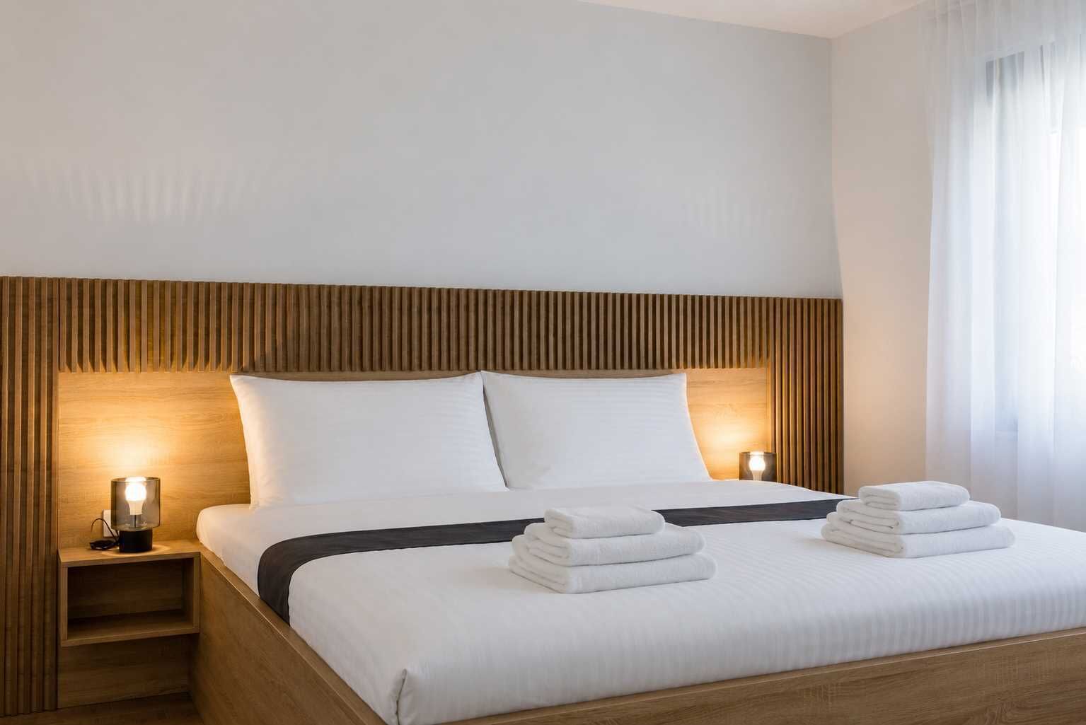 A neatly made bed with white linens and a dark headboard, accompanied by two bedside lamps and a stack of folded towels.