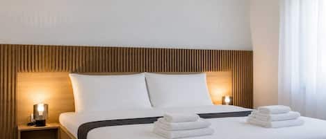 A neatly made bed with white linens and a dark headboard, accompanied by two bedside lamps and a stack of folded towels.