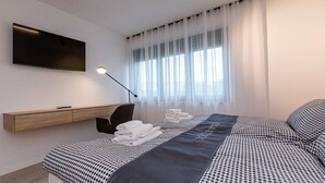 A modern hotel room with a bed, a TV mounted on the wall, a desk, and a window with sheer curtains.