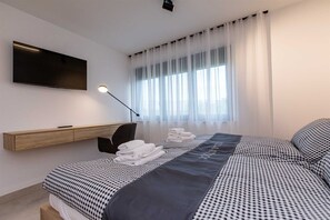 Una habitación de hotel moderna con una cama, un televisor montado en la pared, un escritorio y un ventanal con cortinas translúcidas.