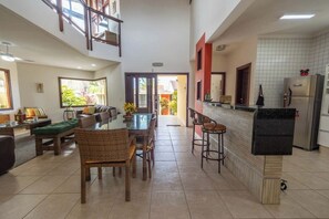 A spacious living area with a dining table, wicker furniture, and a kitchen area with a refrigerator. - Casa com Área Gourmet e Quintal a 200 m da Praia (Itanhaém)