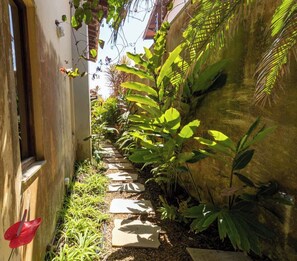 A narrow garden pathway with stepping stones, surrounded by lush green plants and a stone wall. - Casa com Área Gourmet e Quintal a 200 m da Praia (Itanhaém)