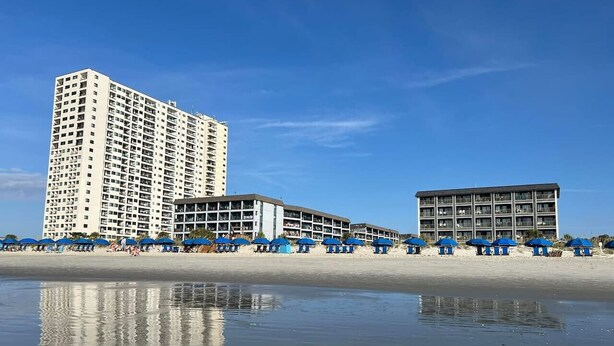 Un hotel frente a la playa con varios pisos, un área de piscina y sombrillas.