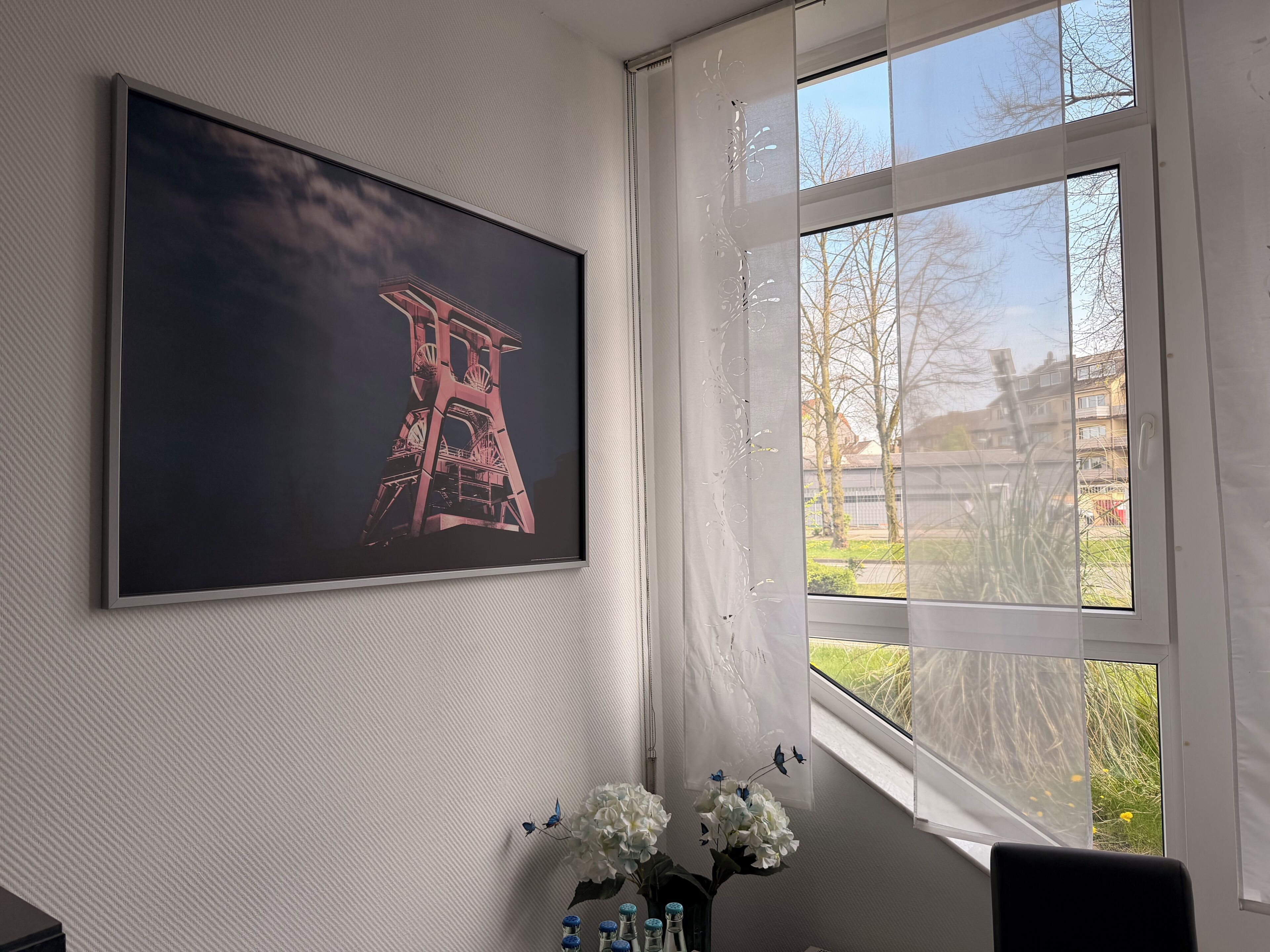 A room with a large framed picture on the wall, a window with a view of buildings, and a vase with flowers on a table.