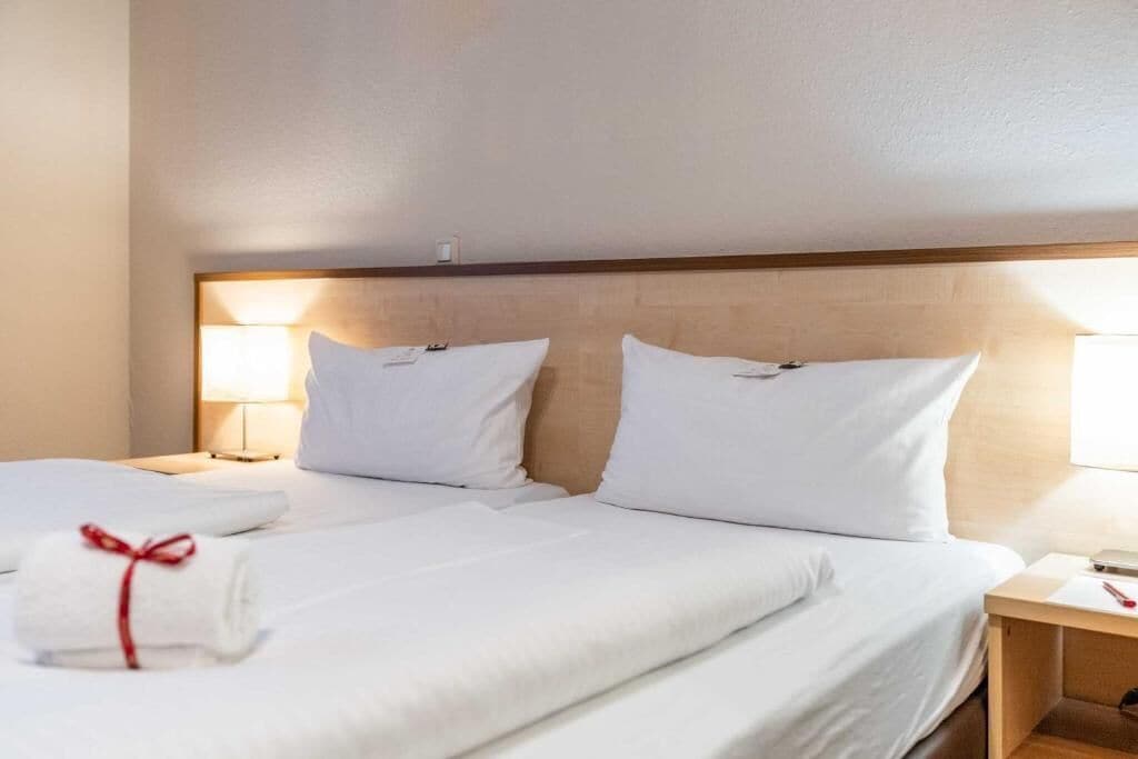 Ein Hotelzimmer mit zwei Betten, weißer Bettwäsche, einem hölzernen Kopfteil, Nachttischlampen und einem ordentlich bezogenen Bett mit einem Handtuch.
