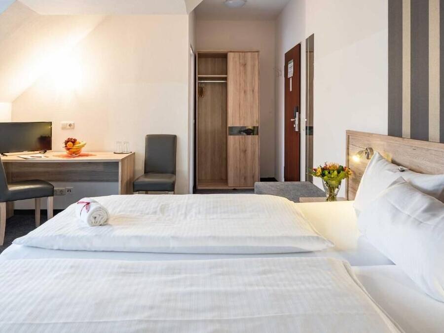 A hotel room with a bed, a desk with a computer, a chair, a wardrobe, and a bedside table with flowers.