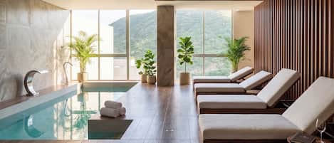 A modern indoor pool area with lounge chairs, a pool, and large windows offering a view of the hills.