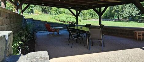 Un patio al aire libre con una mesa y sillas, un muro de piedra y vistas a un área verde con árboles.