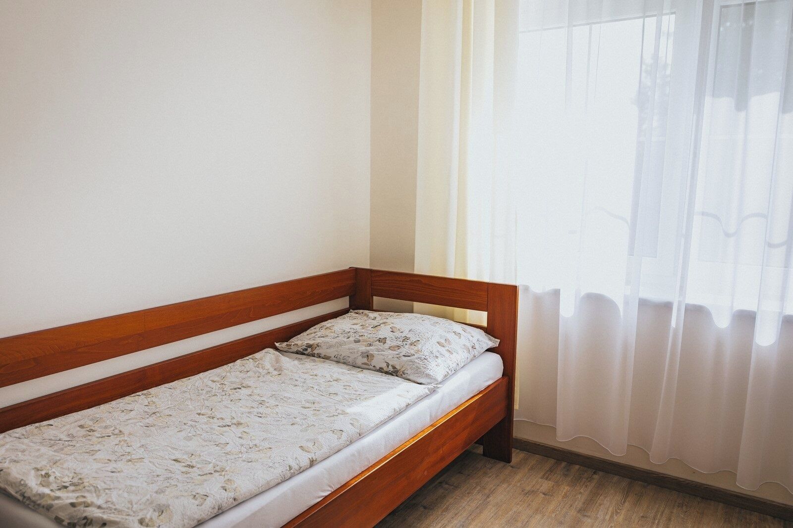 Una cama individual con estructura de madera y ropa de cama floral en una habitación con ventana y cortinas translúcidas.