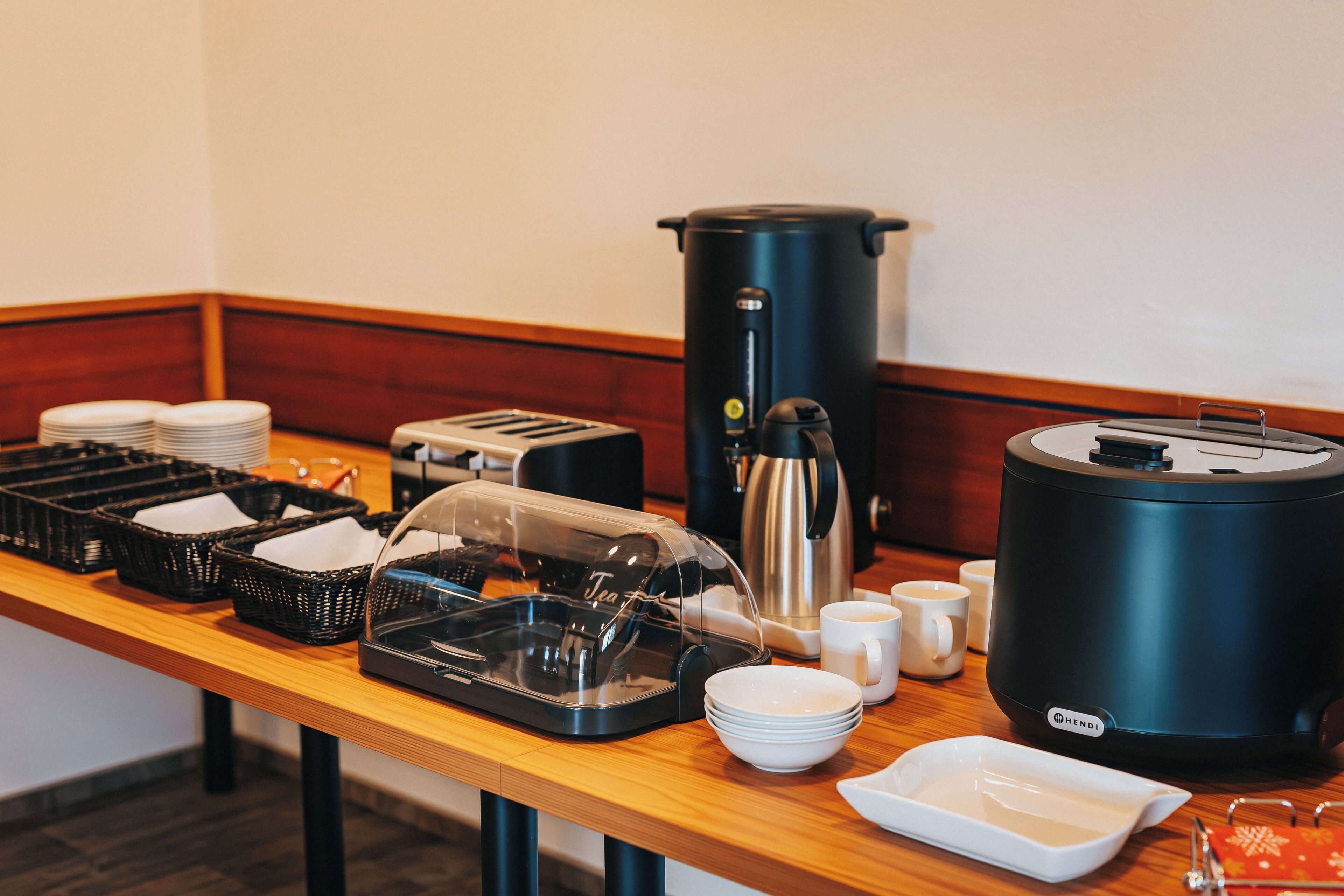 Een buffetopstelling met een koffiezetapparaat, theeservies en stapels borden op een houten tafel.