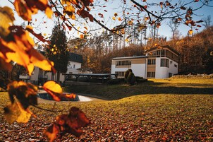 A modern house with a large deck and a swimming pool, surrounded by autumn foliage.