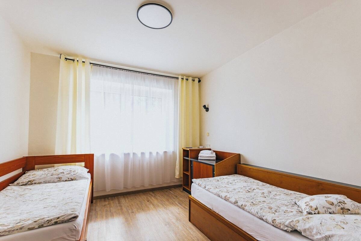 Ένα μικρό δωμάτιο με δύο ξύλινα κρεβάτια, ένα παράθυρο με κουρτίνες, ένα μικρό ράφι και ένα φωτιστικό στην οροφή.