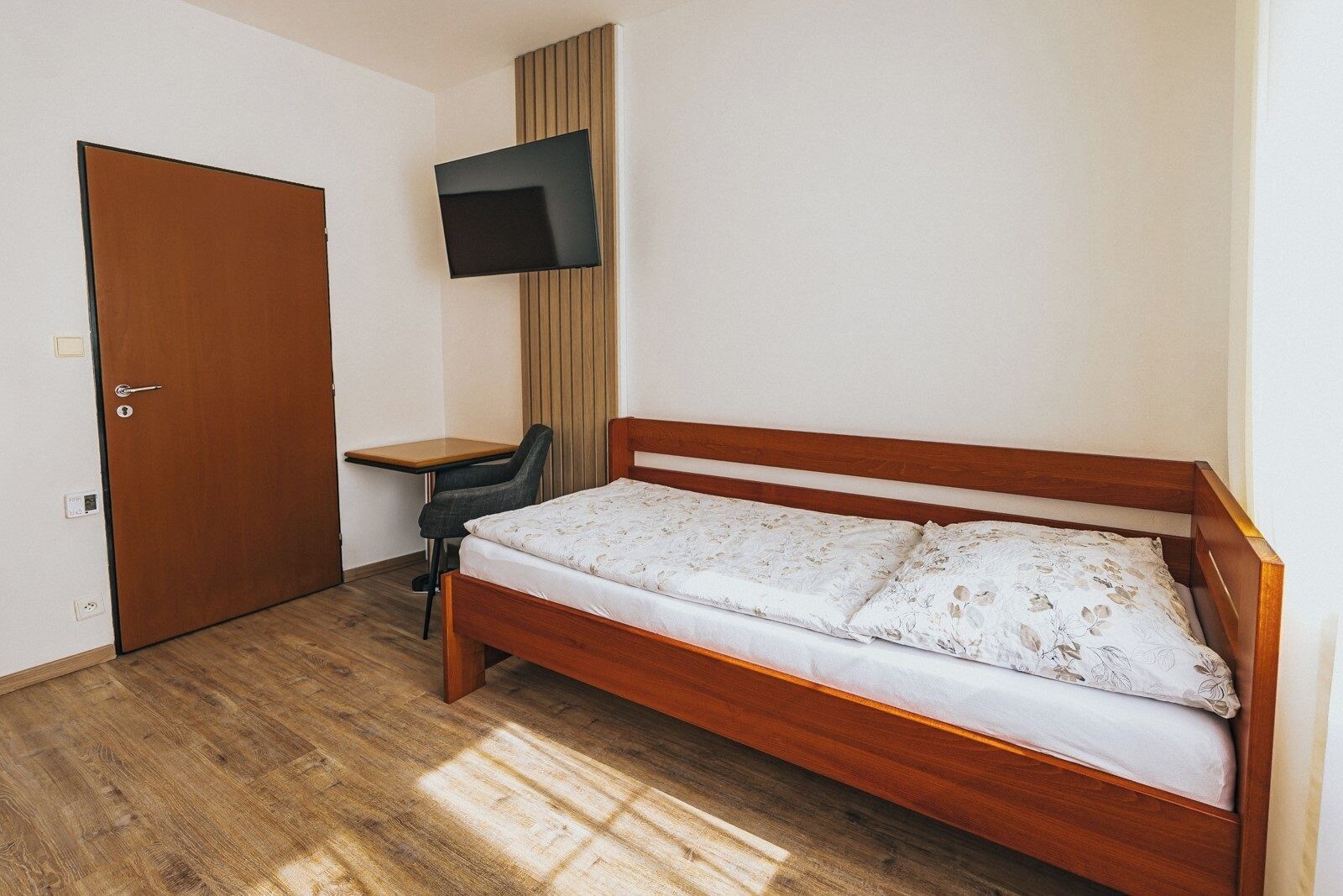 A single bed with a wooden frame and floral bedding, a desk with a chair, a television mounted on the wall, and a door.