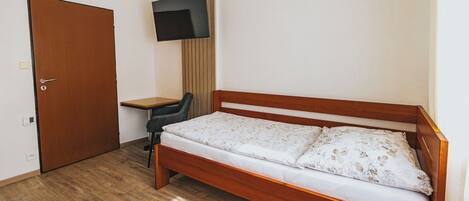 Una cama individual con estructura de madera y ropa de cama floral, un escritorio con silla, un televisor montado en la pared y una puerta.