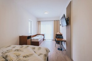 Habitación de hotel compacta con cama, escritorio, silla, televisor montado en la pared y ventana con cortinas.
