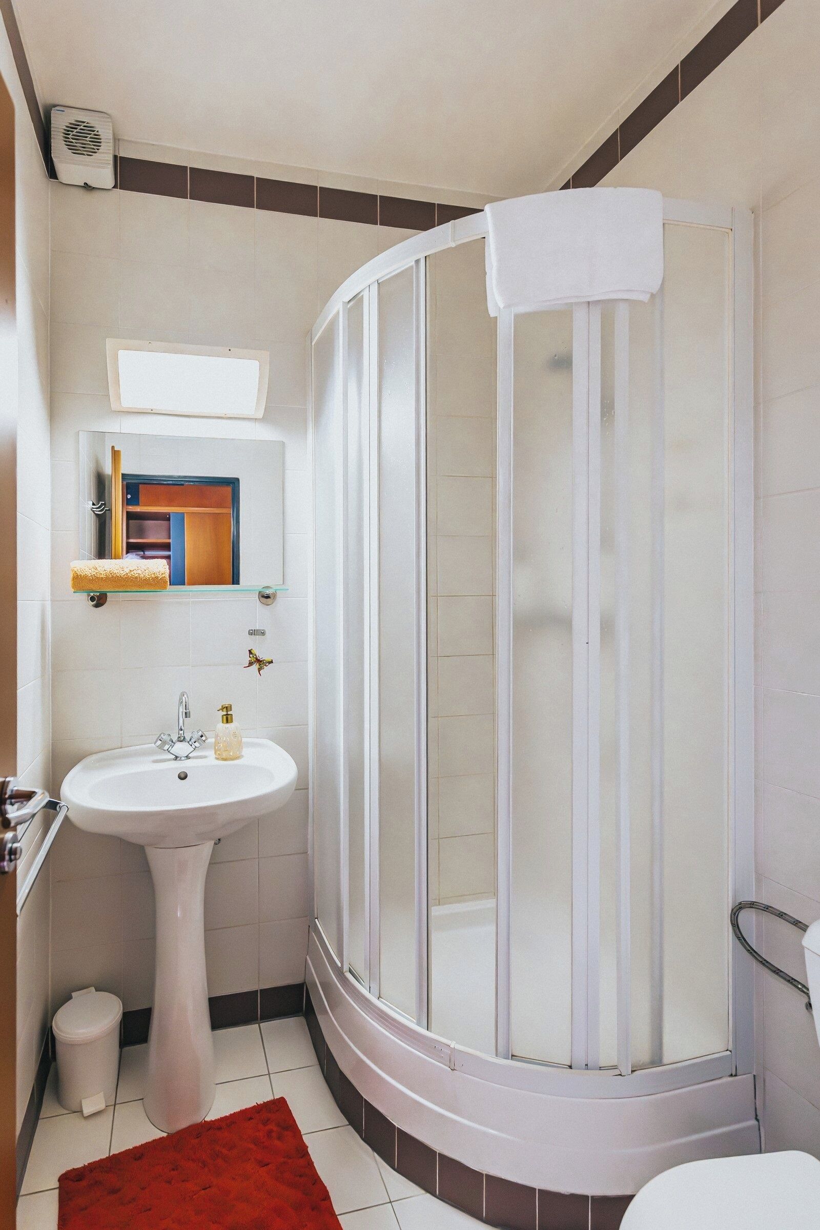 A bathroom with a white sink, a glass shower enclosure, a mirror, and a red rug.