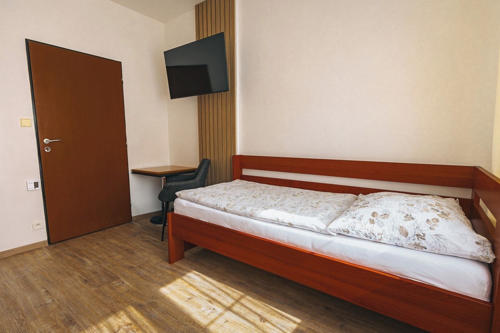 A room with a wooden bed, a desk, a chair, a TV mounted on the wall, and a door.
