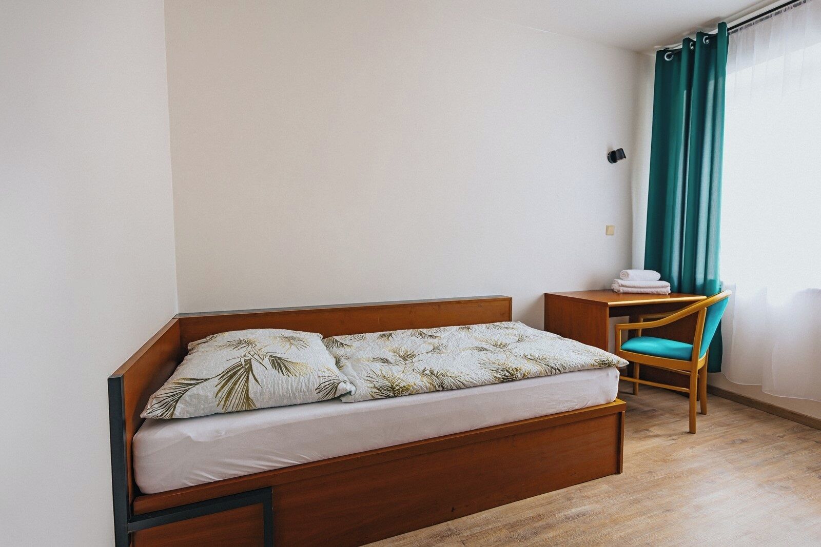 A single bed with a wooden frame, a small bedside table, a chair, and a window with curtains.