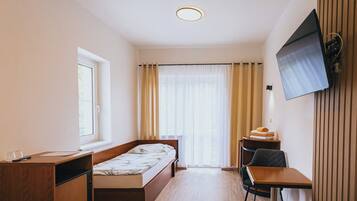Ein kleines Hotelzimmer mit Bett, Schreibtisch, Stuhl, Wandfernseher und einem fenster mit Vorhängen.